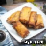 Side shot of multiple egg rolls arranged on a white square plate on a wooden surface with a blue striped cloth and a dipping sauce in a small white bowl on the side.
