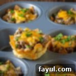 Side shot of a wonton cheeseburger cup sitting on a the flat part of a cupcake sheet with more in the background.