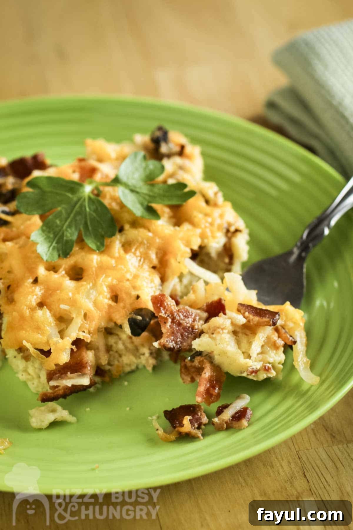 A delectable slice of Bacon and Hash Brown Breakfast Casserole on a vibrant green plate, with a fork poised to take a bite. A green cloth napkins sits invitingly beside it.