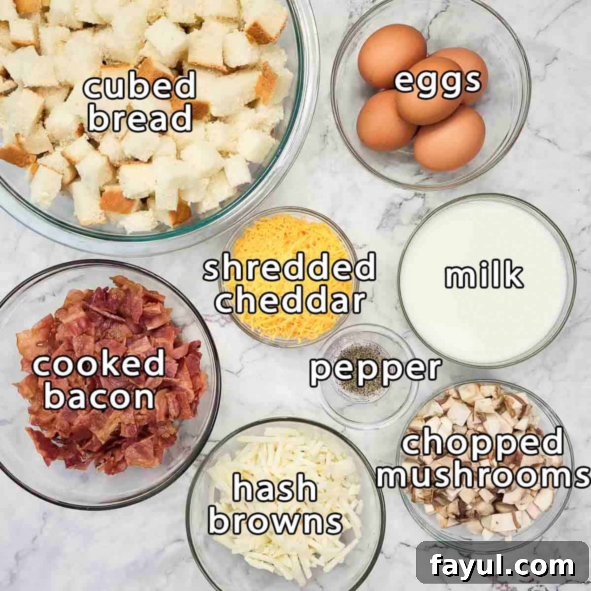An overhead shot showcasing all the fresh ingredients for the breakfast casserole: cubed bread, fresh eggs, shredded cheddar cheese, milk, crispy cooked bacon, frozen hash browns, chopped mushrooms, and black pepper.