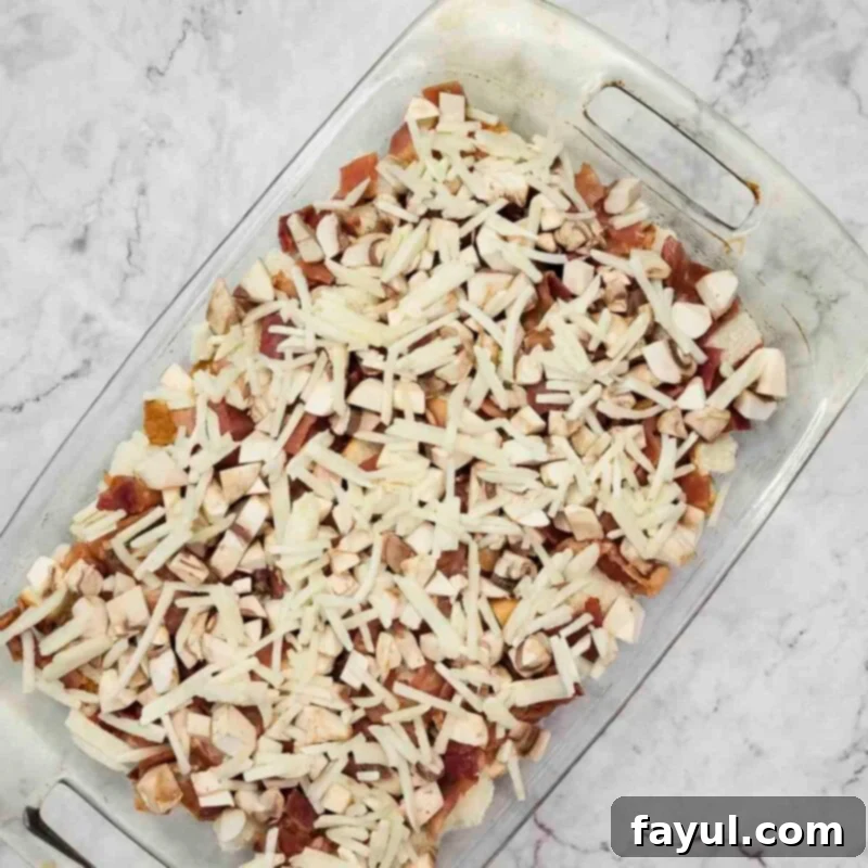 Overhead view of the fourth step: spreading frozen hash browns evenly over the bacon and mushroom layers in the casserole dish.
