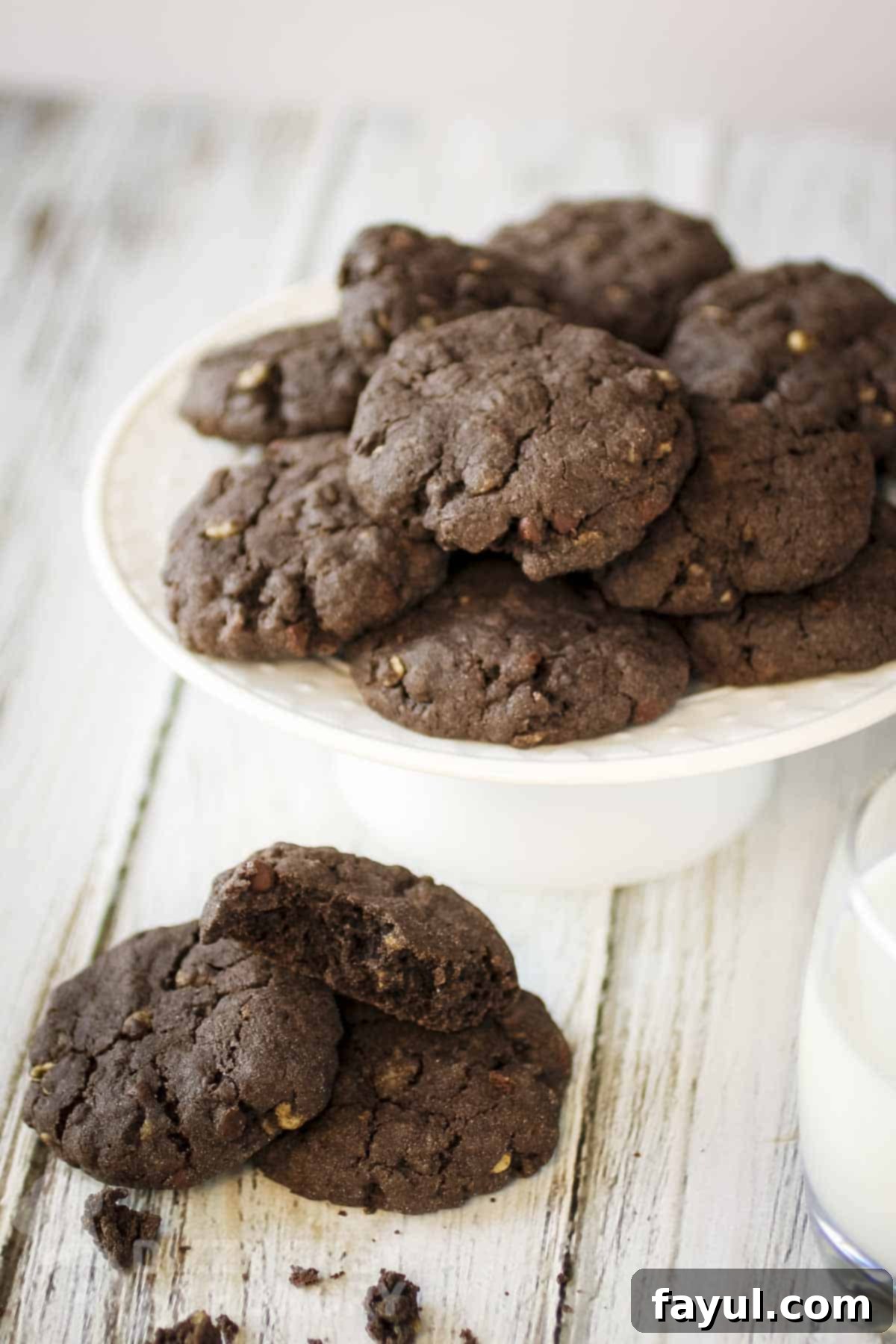 Side shot of multiple chocolate cookies stacked on a white serving platter and more on a wooden surface below.