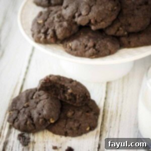 Side shot of multiple chocolate cookies stacked on a white serving platter and more on a wooden surface below.