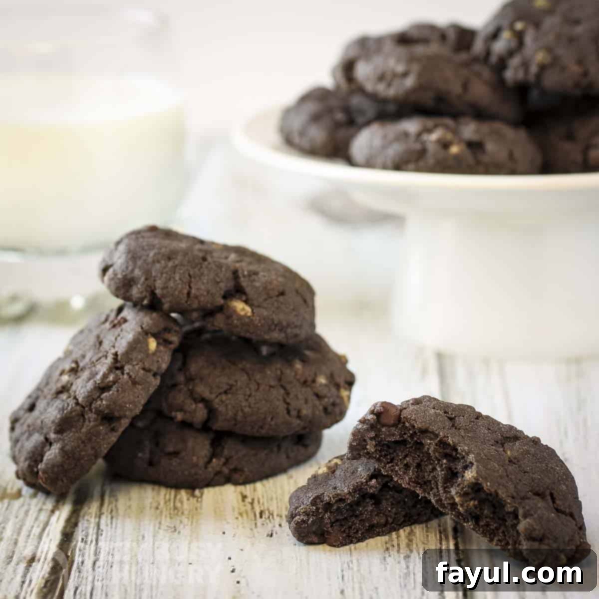 Side shot of multiple chocolate cookies stacked on a white serving platter and more on a wooden surface below, one broken in half.