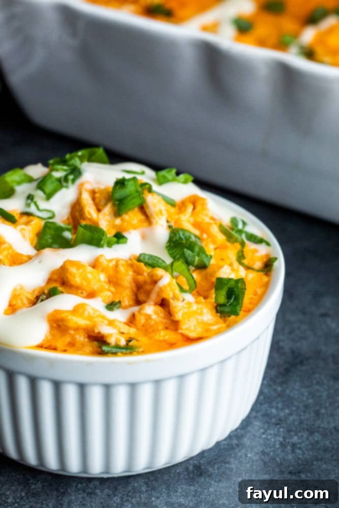 Close-up shot of baked buffalo chicken dip in a white bowl, topped with a drizzle of ranch and chopped green onions.