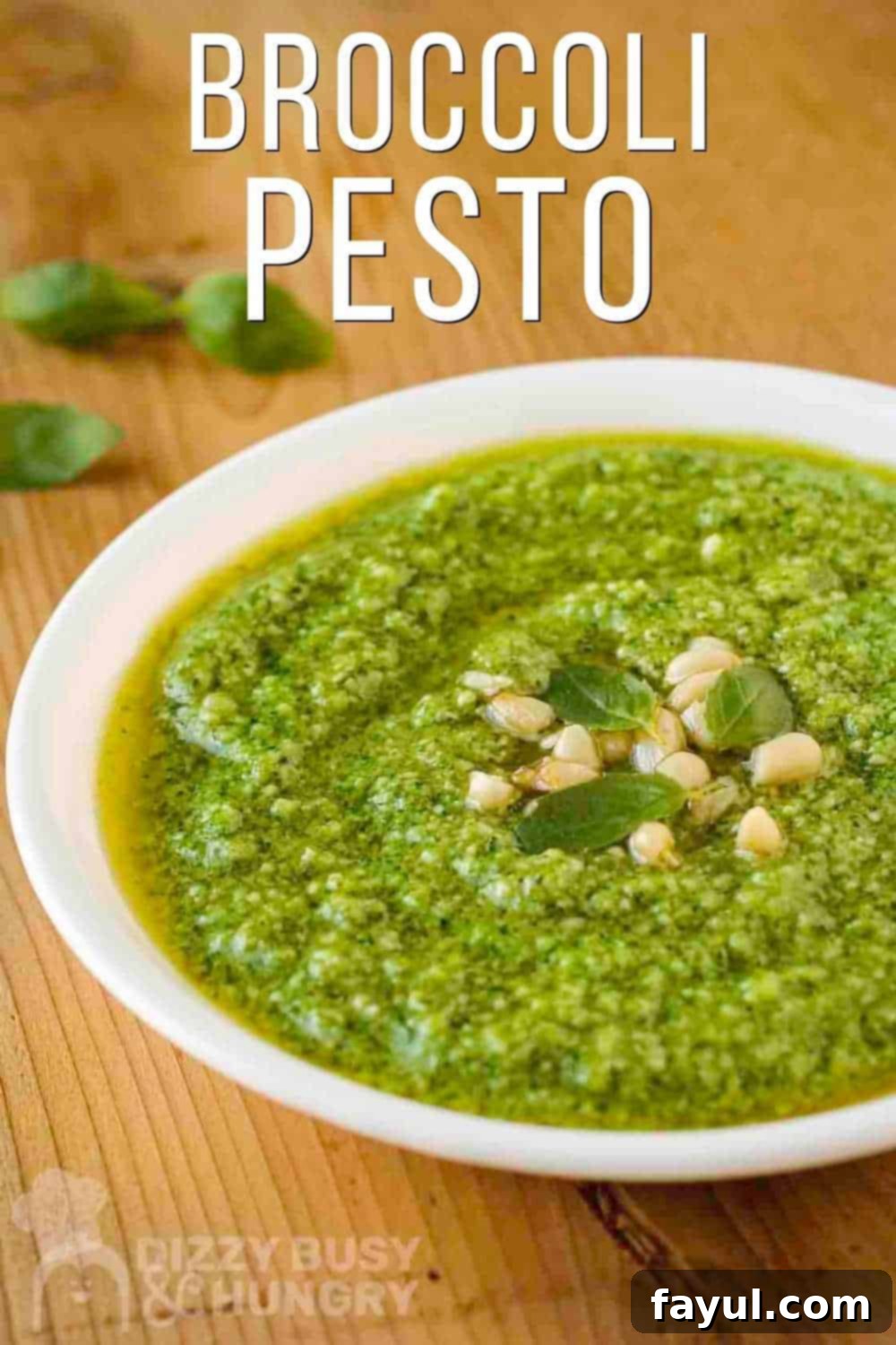 Side view of vibrant green broccoli pesto in a white bowl garnished with fresh basil leaves and golden pine nuts, with more basil on a wooden cutting board in the background.
