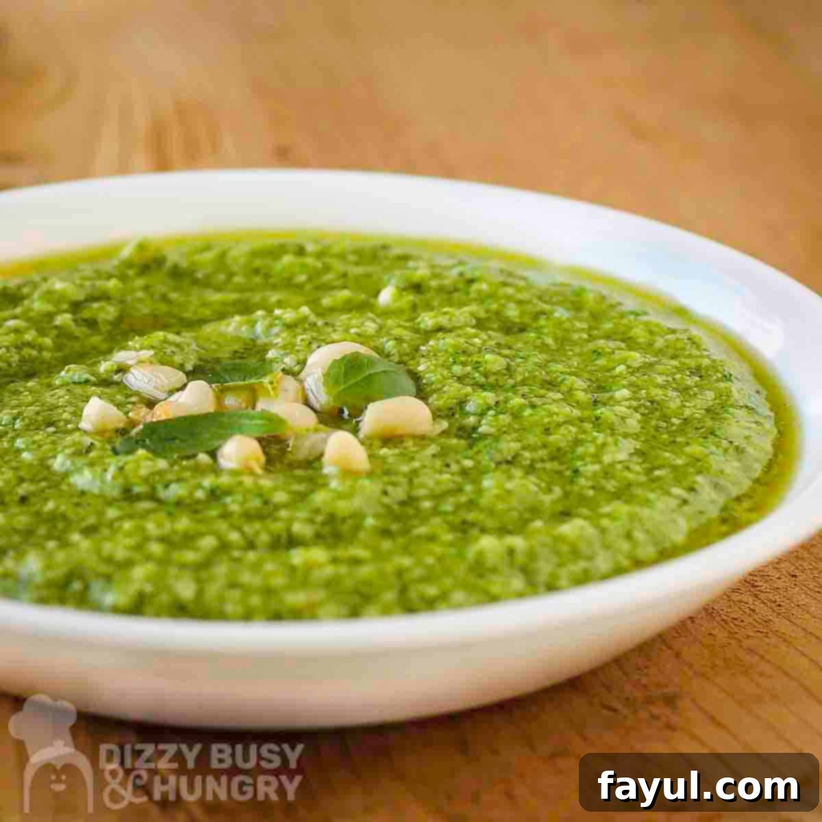 Side close-up view of vibrant green broccoli pesto in a white bowl garnished with fresh basil leaves and golden pine nuts, with more basil on a wooden cutting board in the background, showcasing its rich texture.