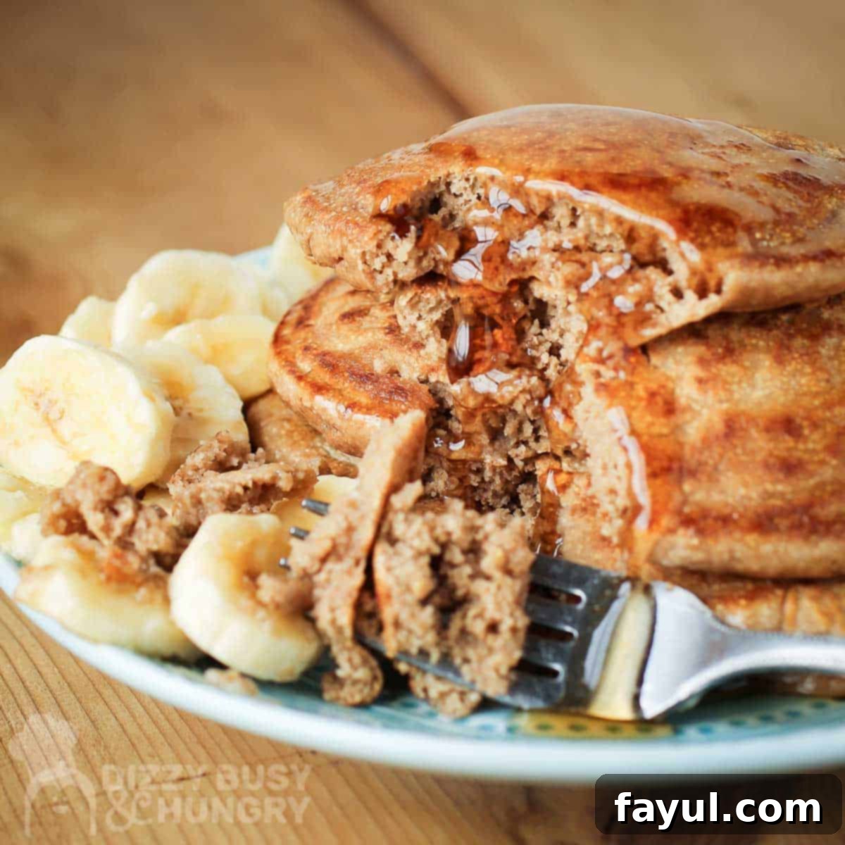Side shot of a fork taking pieces out of multiple stacked pancakes with a syrup drizzle and sliced bananas on the side on a light blue plate.