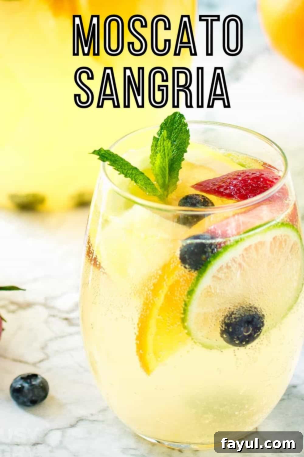 Side shot of a glass wine tumbler with sangria garnished with mint and fruits on a marble surface with a pitcher of it in the background.