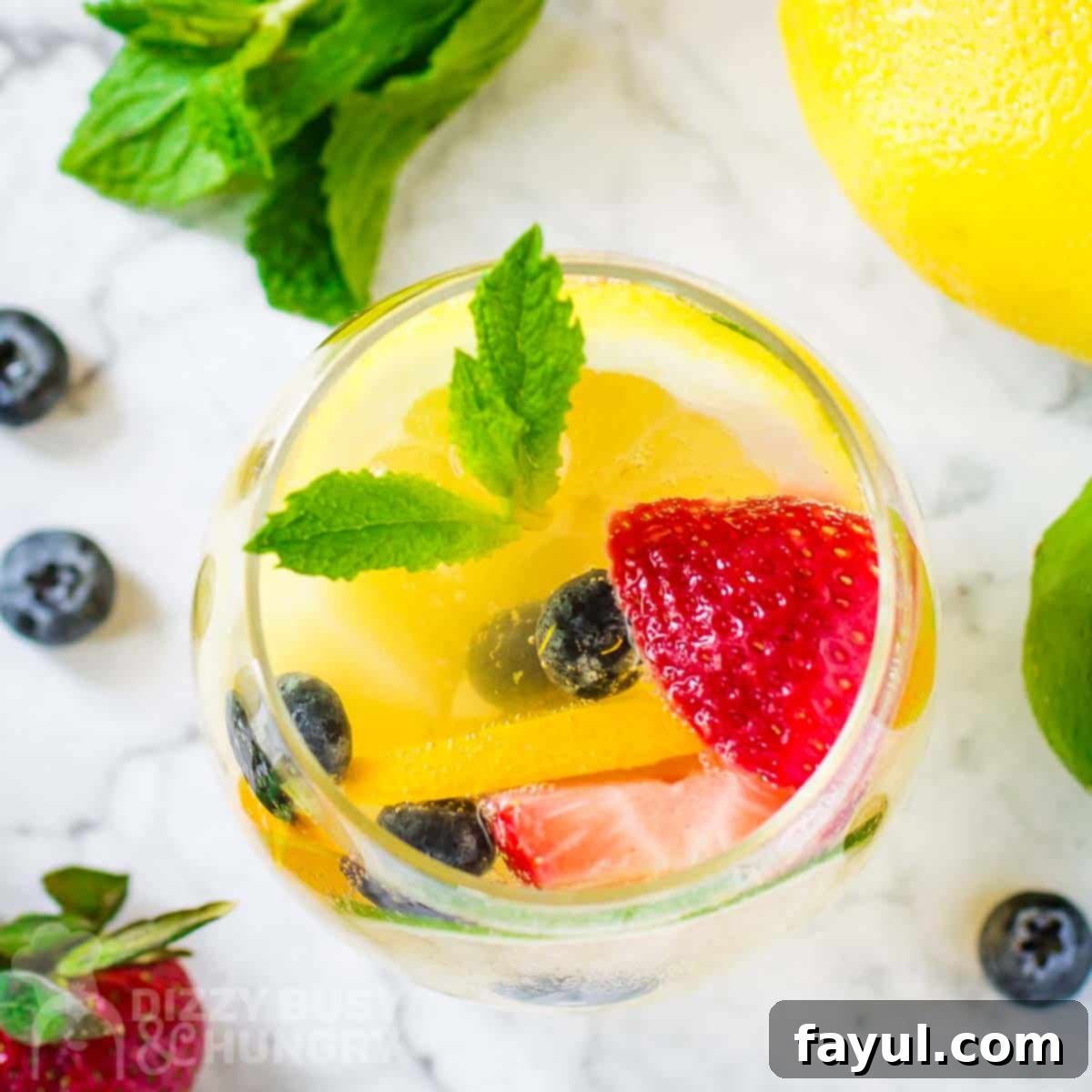 Overhead shot of a glass wine tumbler with sangria garnished with mint and fruits on a marble surface with fruits scattered around.