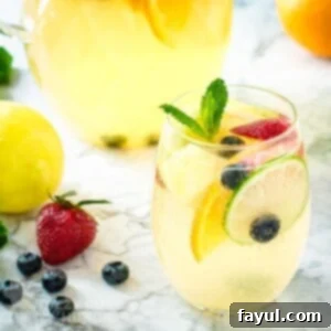 Side shot of a glass wine tumbler with sangria garnished with mint and fruits on a marble surface with a pitcher of it in the background and fruits scattered on the side.