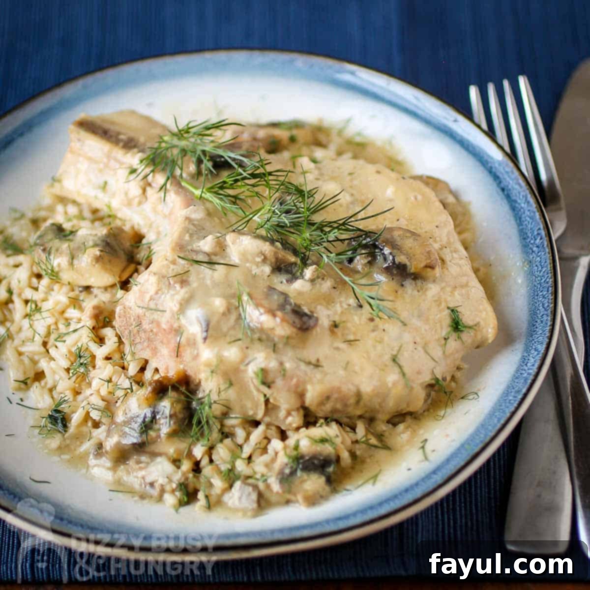 Side close up view of a tender pork chop on top of mushroom soup garnished with fresh herbs on a blue and white plate with silverware on the side on a dark blue placemat. A comforting crock pot meal.