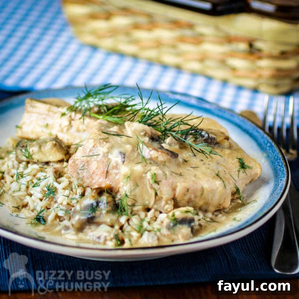 Side angled view of a pork chop on top of mushroom soup garnished with herbs on a blue and white plate with silverware on the side on a dark blue placemat and a basket in the background. A perfectly cooked slow cooker mushroom pork chop.