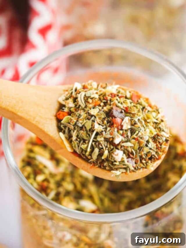 Close up shot of a wooden spoon holding a scoop of vibrant pizza spices over a clear glass jar full of them, with a red cloth in the soft background, highlighting the aromatic blend.
