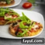 Side shot of two mini Caprese pizzas elegantly arranged on a white plate, with a fresh tomato and greens blurred in the background.