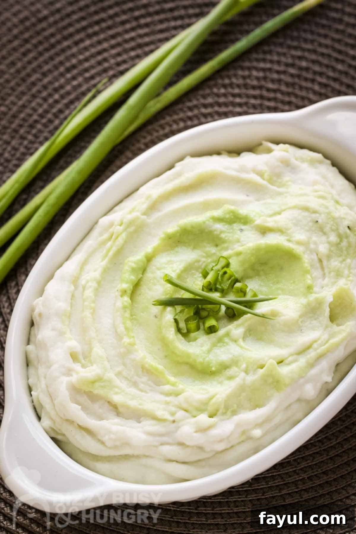Overhead close up shot of wasabi mashed potatoes in a white baking dish garnished with green onions and chives with more green onions in the background.