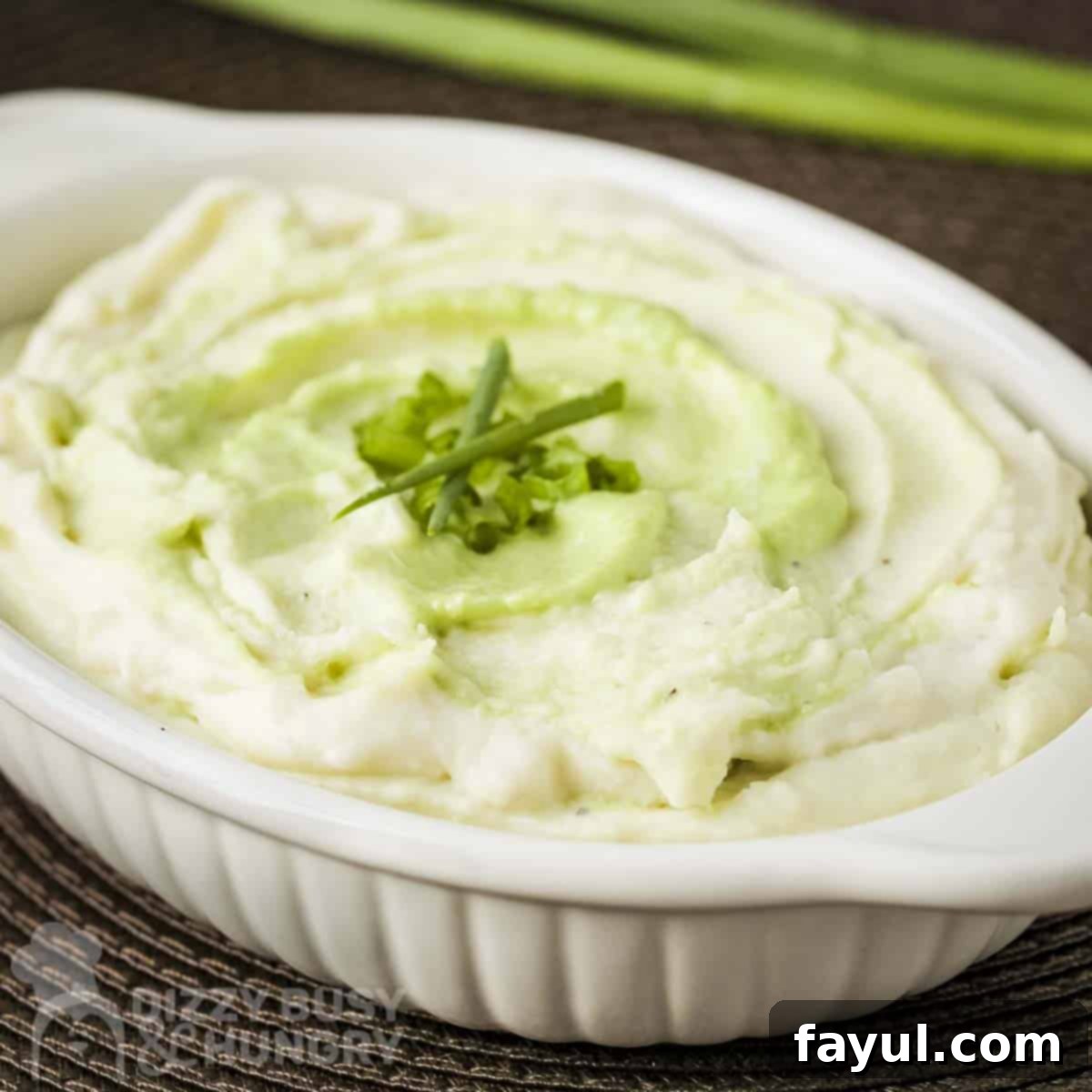 Side close up shot of wasabi mashed potatoes in a white baking dish garnished with green onions and chives with more green onions in the background.