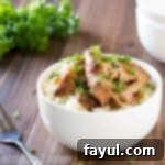 Side shot of crock pot chicken thighs in a white bowl on a wooden surface with herbs and a fork in the background.