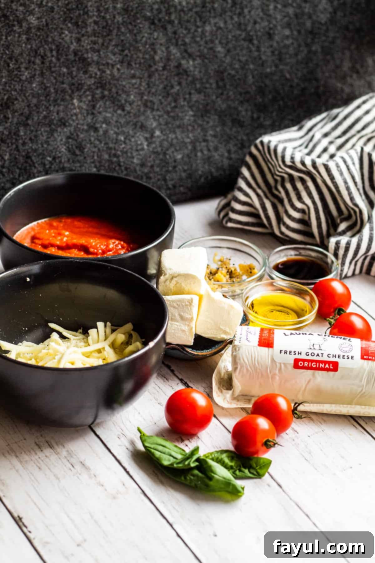 Goat cheese ingredients on a white counter laid out with black background, including blocks of cheese, tomatoes, basil, and a jar of marinara.
