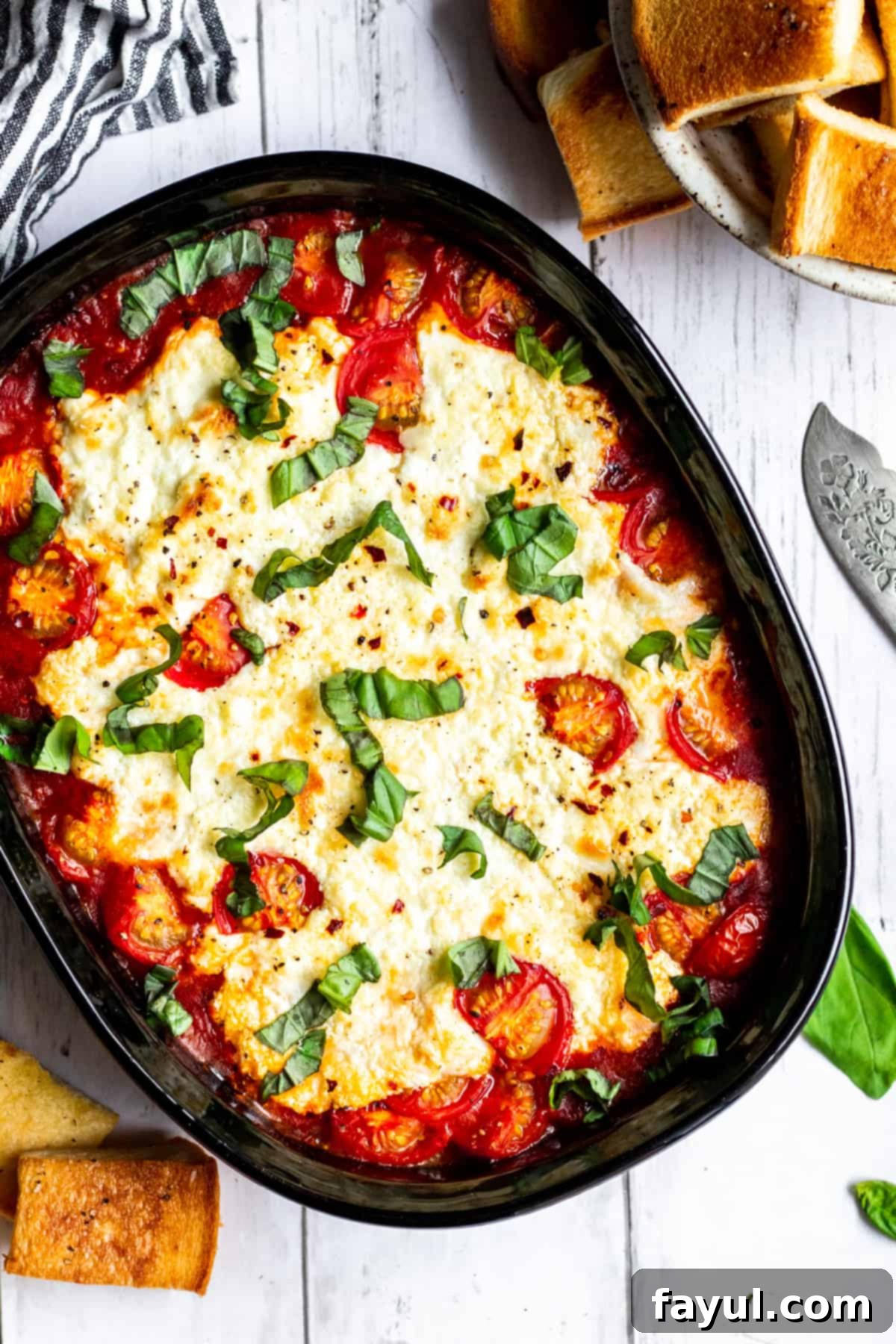 Wide overhead shot of baked goat cheese dip on a white counter with toasted bread slices and a kitchen towel surrounding it, ready for serving.
