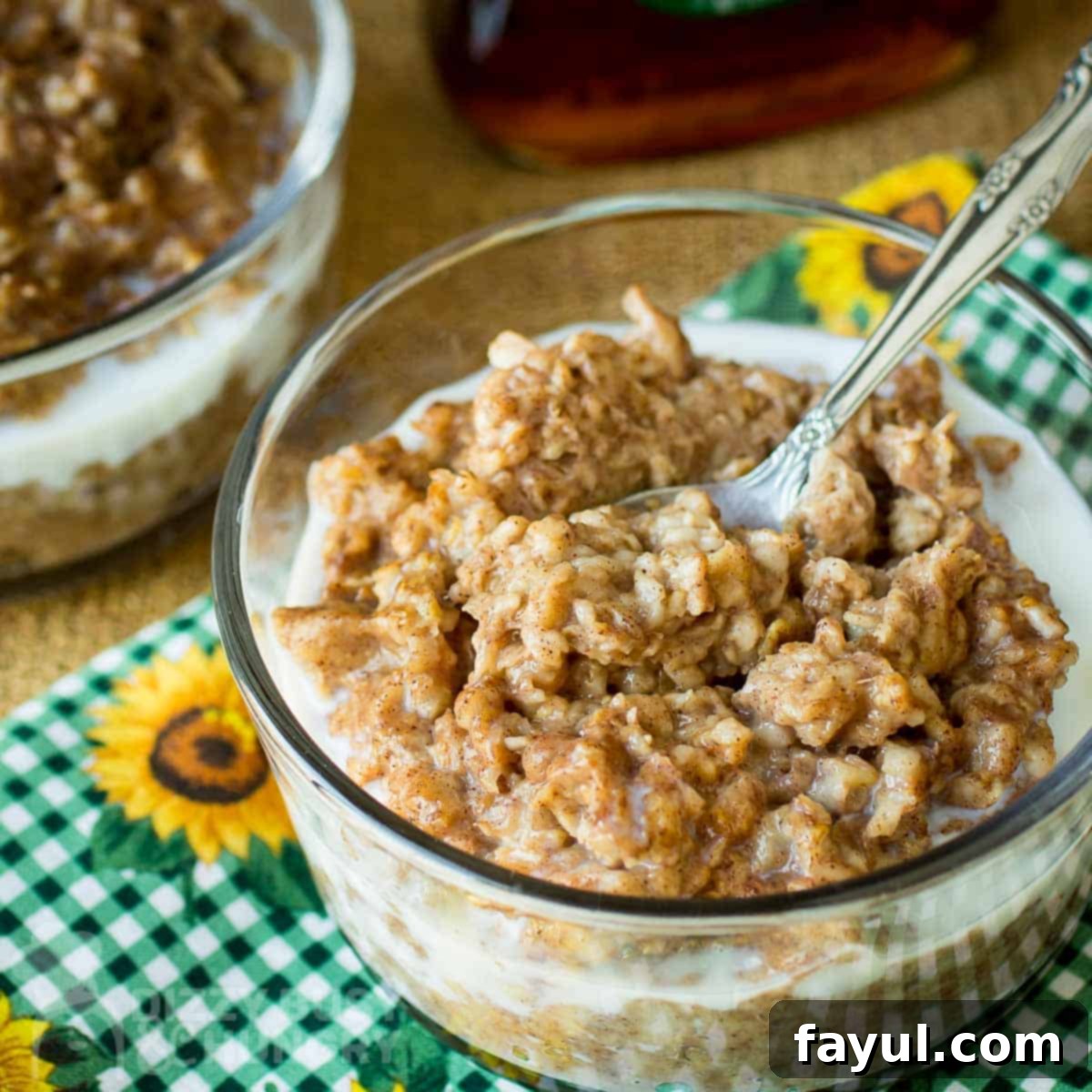Side angled shot of oatmeal in a clear bowl with a spoon on a green checkered cloth with sunflowers, showcasing the creamy texture.