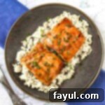 Close up shot of glistening teriyaki glazed salmon served over white rice, garnished with fresh chives, presented on a rustic brown plate with a blurred blue background.