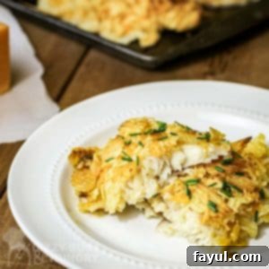 Side view of potato crusted cod on a white plate on a wooden surface with more cod on a baking sheet in the background, showcasing the crispy golden crust.