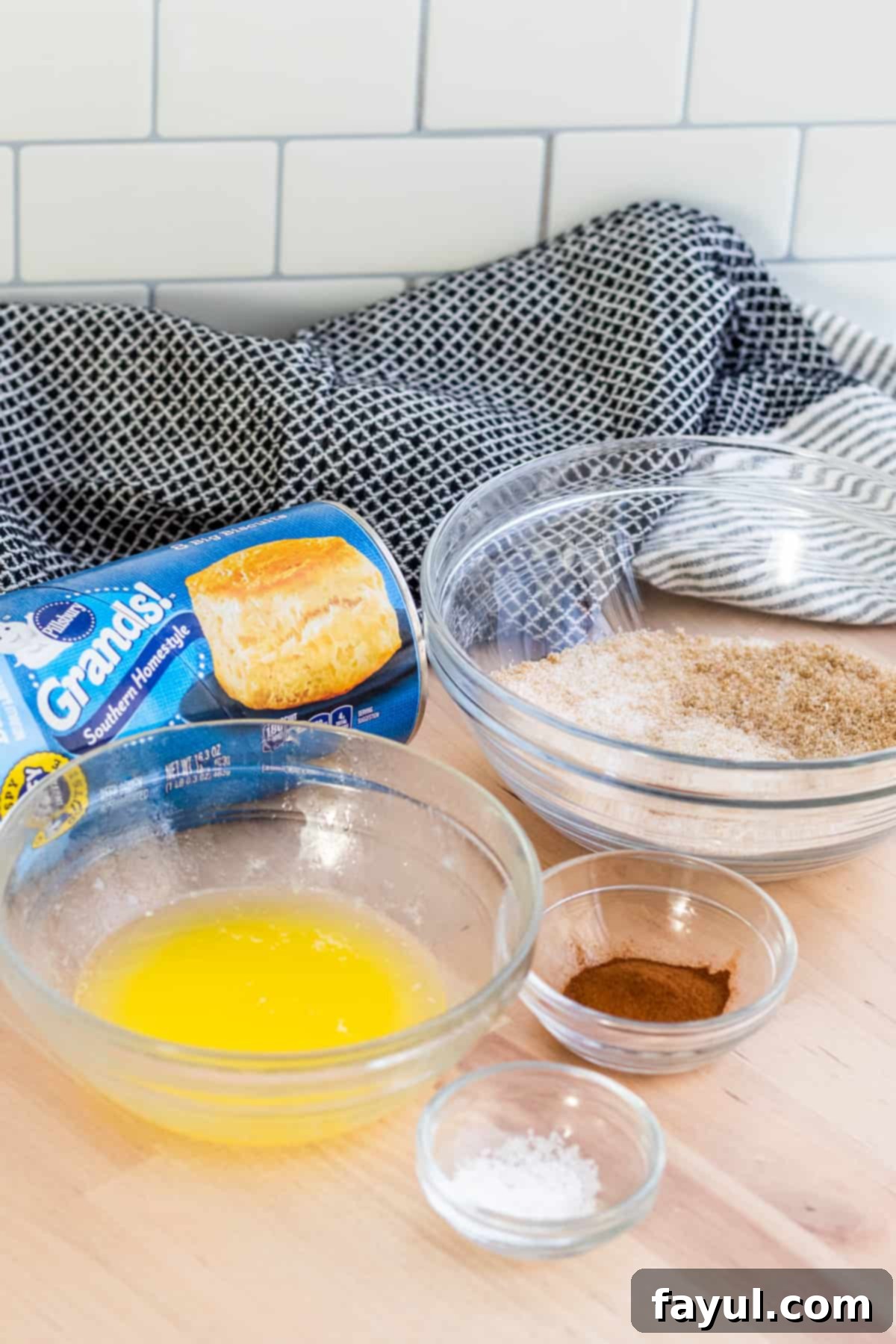 Easy Air Fryer Cinnamon Sugar Biscuit Donuts 3 Close-up shot of donut ingredients neatly arranged in glass bowls on a rustic wooden table, including canned biscuits, butter, and cinnamon sugar mix.
