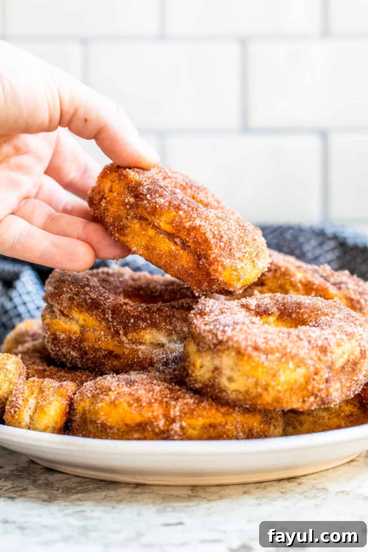 Easy Air Fryer Cinnamon Sugar Biscuit Donuts 6 A hand reaching into a pile of warm, coated air fryer donuts on a white plate, ready to pick one up.