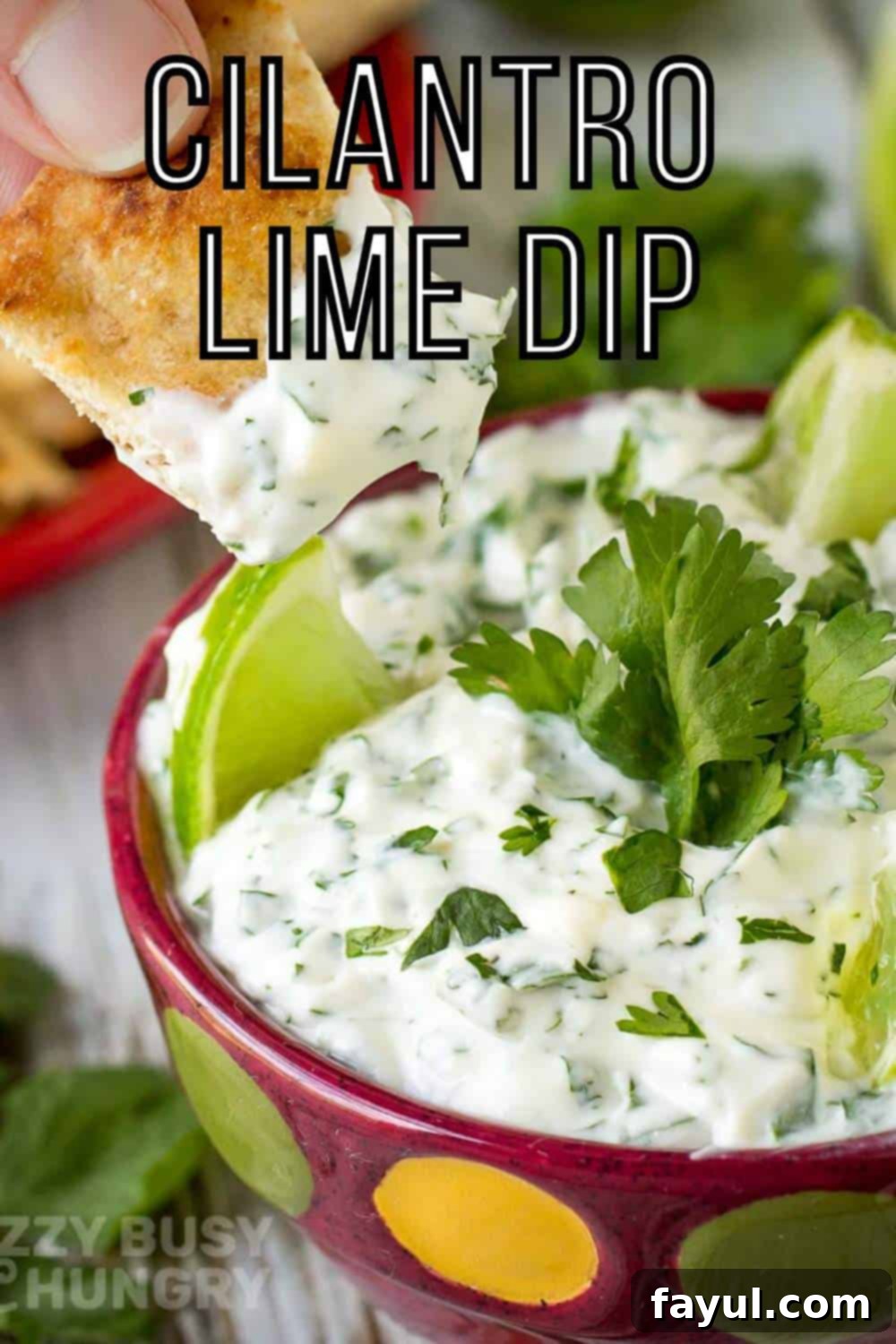 Close up shot of a chip being dipped into cilantro lime dip with sliced limes and cilantro in the background.
