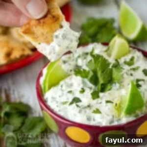 Close up shot of a chip being dipped into cilantro lime dip with sliced limes and cilantro in the background.