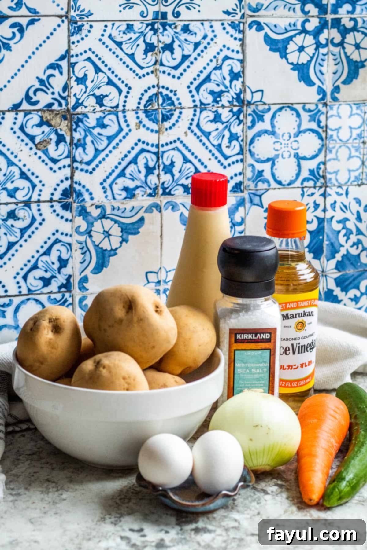 Ingredients for Japanese potato salad laid out on a white table in bowls and small dishes, including potatoes, carrots, cucumbers, eggs, and mayonnaise.
