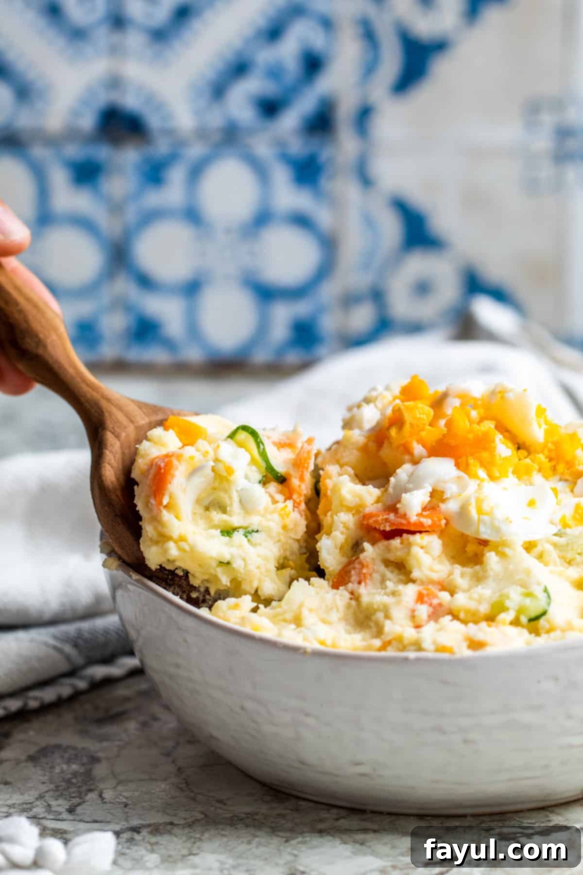 Hand using a wooden spoon to scoop out a portion of Japanese potato salad from a white bowl.