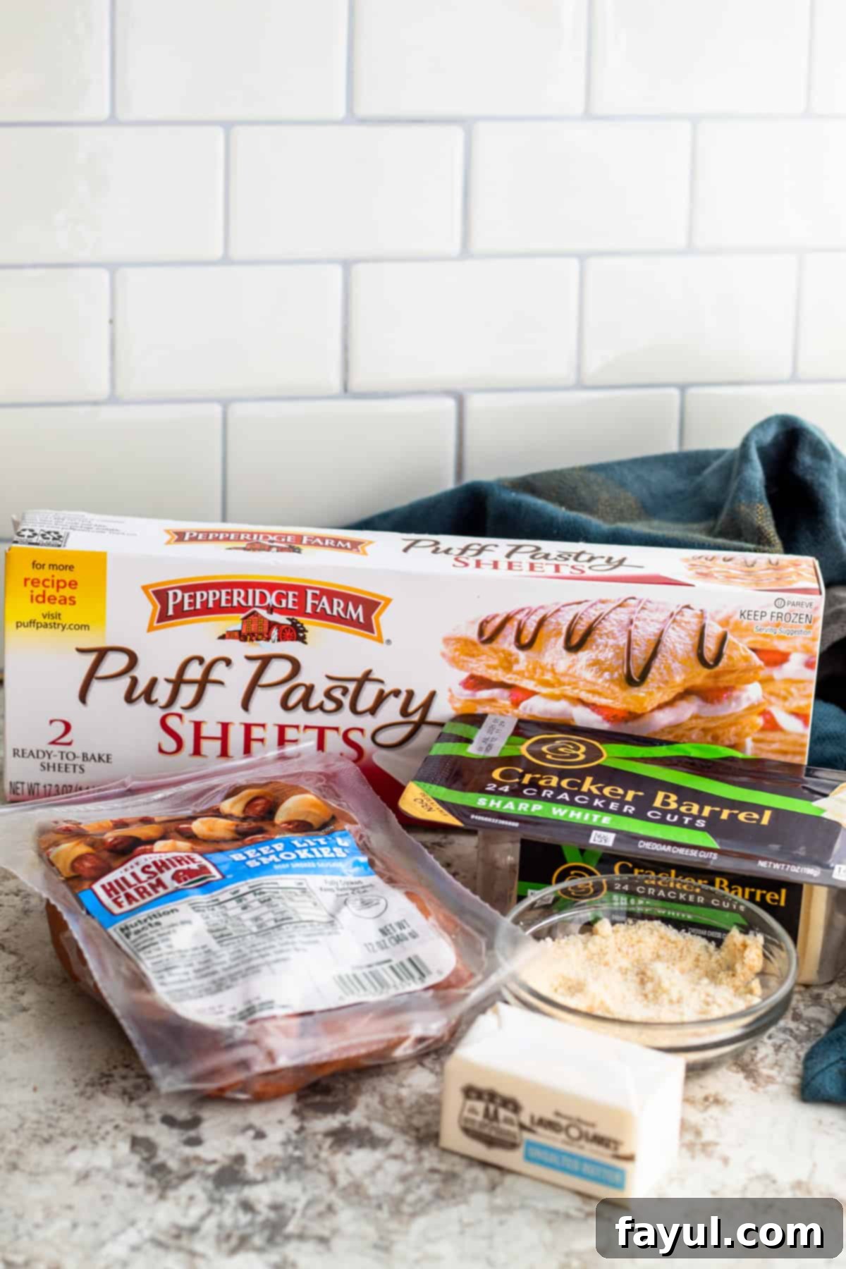 Flaky Cheesy Pigs in a Blanket 3 All the ingredients for Cheesy Pigs in a Blanket laid out neatly on a white kitchen counter, including puff pastry, lil' smokies, white cheddar cheese slices, melted butter, and garlic parmesan seasoning mixture.