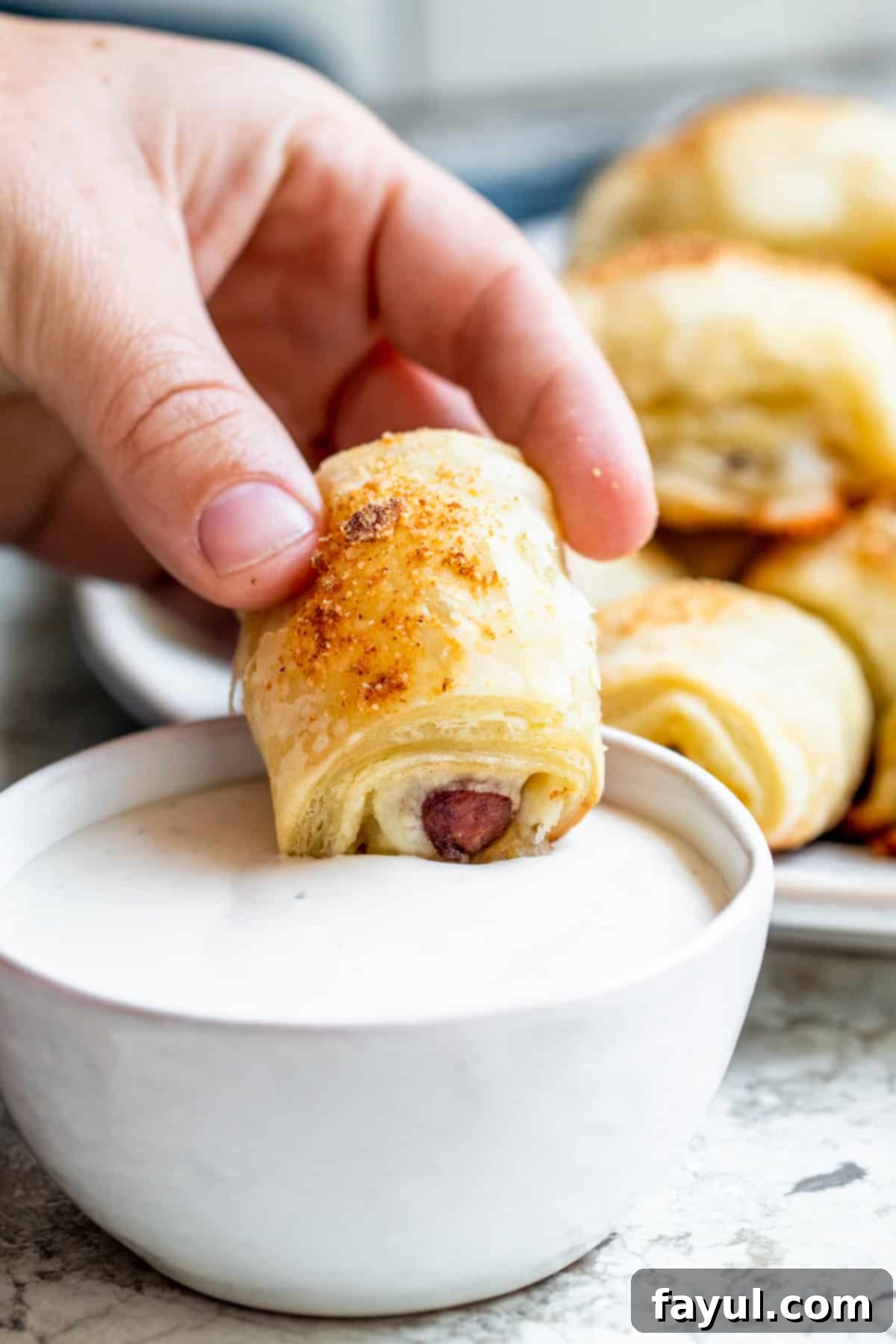 Flaky Cheesy Pigs in a Blanket 5 A hand dipping a freshly baked mini crescent dog, or pig in a blanket, into a small white bowl of creamy ranch dressing, highlighting the perfect pairing.