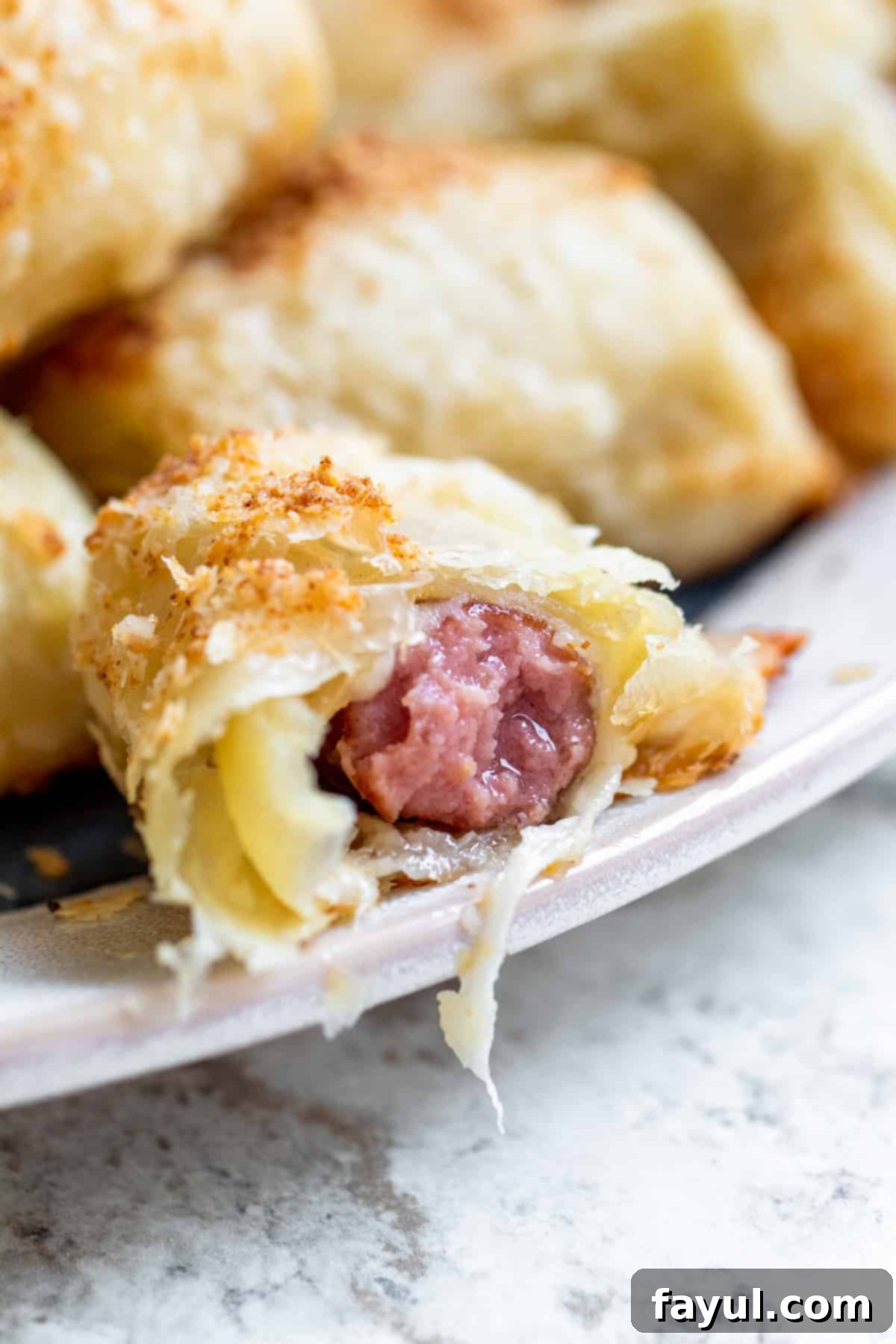 Flaky Cheesy Pigs in a Blanket 6 A close-up, straight-on shot of a cheesy pig in a blanket on a plate with a bite taken out, revealing the melted cheese and sausage inside, showcasing its deliciousness.