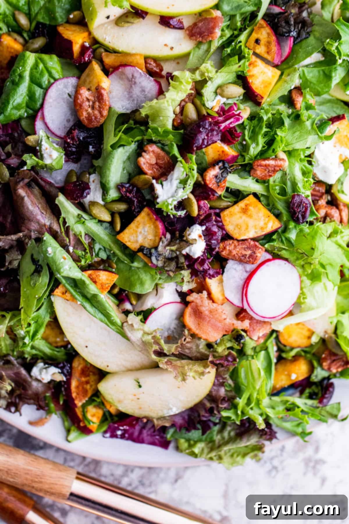 An overhead shot of a Fall Harvest Salad artfully arranged on a white plate, served on a clean white kitchen counter. Elegant gold serving spoons rest beside the plate, emphasizing the salad's readiness for a special meal or gathering.