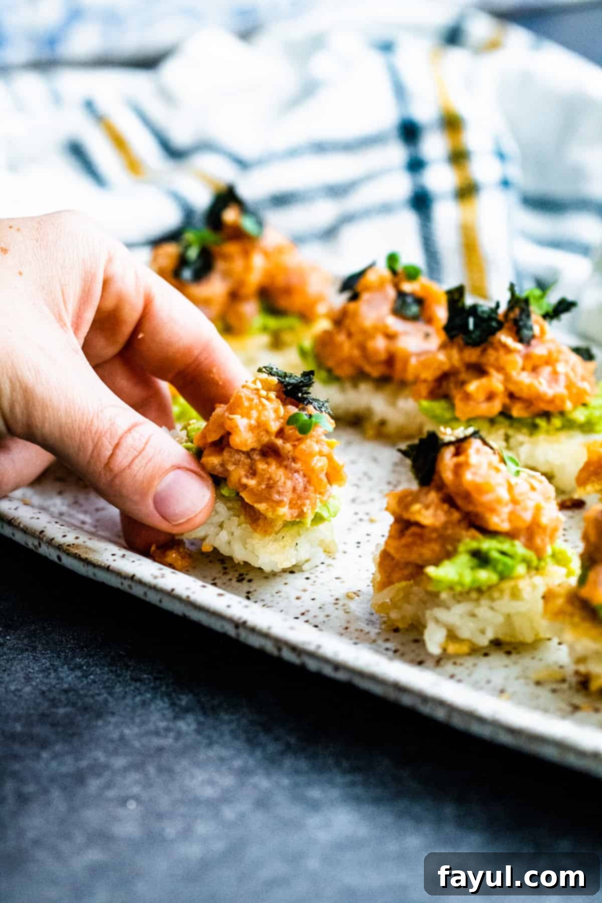 A hand gently picking up a piece of Spicy Tuna Crispy Rice Sushi from a neatly arranged platter, showcasing its appeal.