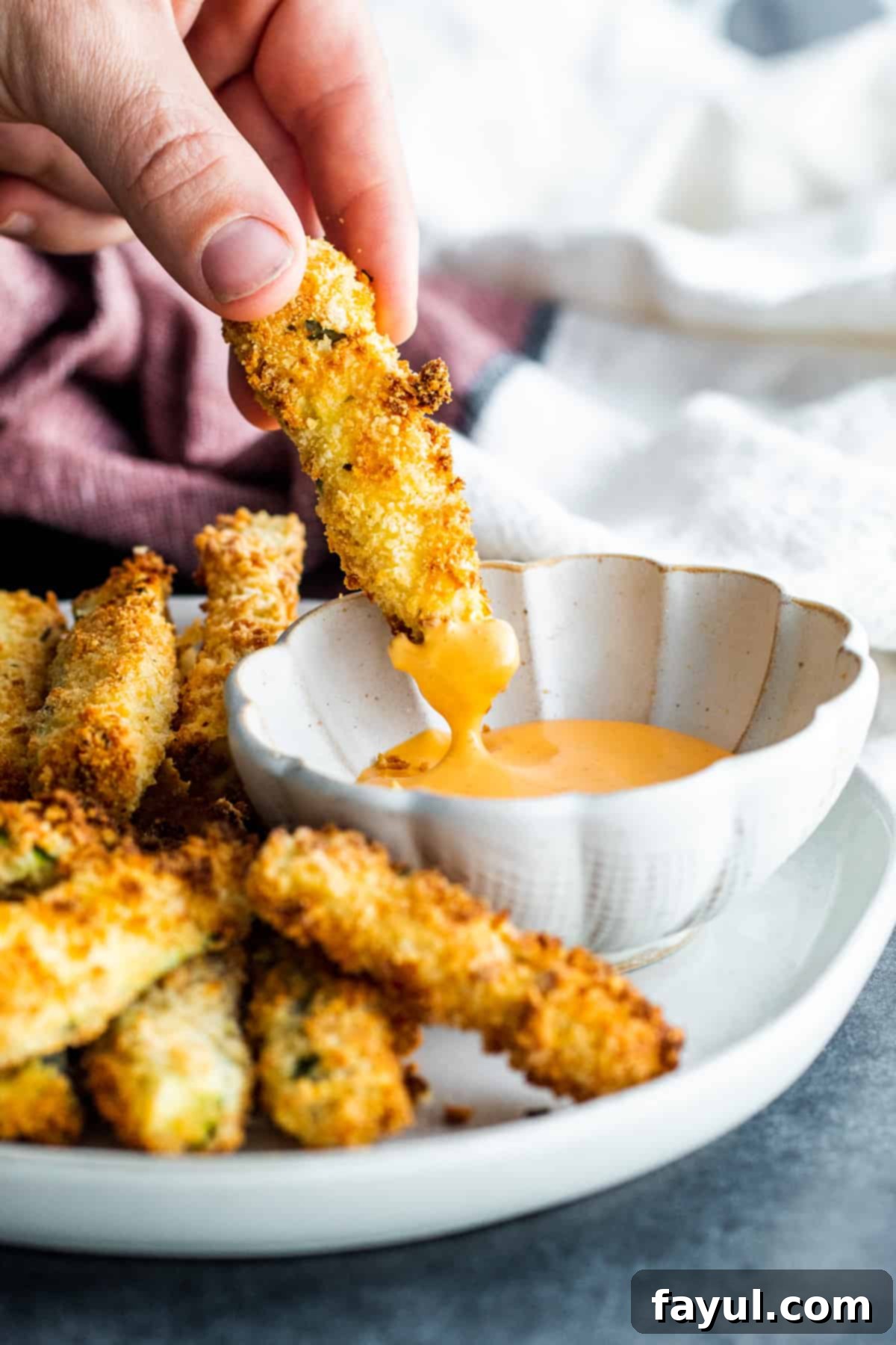 Crispy Garlic Parmesan Air Fryer Zucchini Fries 8 A hand dipping a golden crispy air-fried zucchini fry into a small white bowl of creamy, savory sauce.