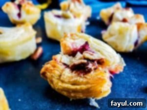 A close-up, straight-on shot of a single blackberry puff pastry bite, showcasing its luscious blackberry syrup filling and golden pastry, resting on a blue kitchen counter. Other bites and a blue and yellow towel are blurred in the background.