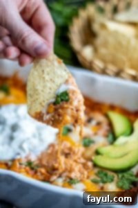 The BEST Baked Chicken Enchilada Dip recipe! This Mexican inspired appetizer is super easy and cheesy and filled with beans, corn and tons of flavor. Top with avocado, sour cream and cilantro for the perfect finish! A hand dipping a tortilla chip into the warm, cheesy chicken enchilada dip. A blurred basket of chips is in the background.