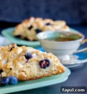 These Blueberry White Chocolate Scones with Lemon Glaze are the perfect sweet breakfast or afternoon tea treat! This easy dessert recipe will be your new family favorite! #scones #breakfast #dessert #blueberry #recipe Strait on shot of scone on a teal plate with second blurred in the background with a white tea cup.