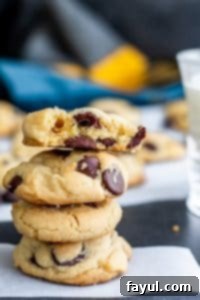 A stack of golden-brown salted chocolate chip cookies on parchment paper, with more freshly baked cookies blurred in the background, showcasing their crisp edges and soft centers.