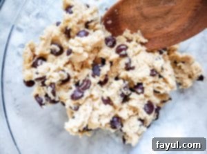 Raw cookie dough in a clear glass bowl with a wooden spoon resting inside, showing the rich texture and visible chocolate chips, ready for baking.