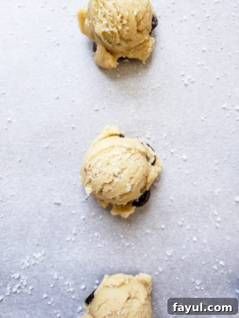 An overhead shot of three perfectly scooped cookie dough balls arranged in a neat line on parchment paper, each topped with a sprinkle of sea salt, ready to be baked into delicious salted chocolate chip cookies.