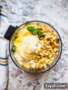Prep image for pesto sauce showing raw ingredients in a food processor on a white counter.