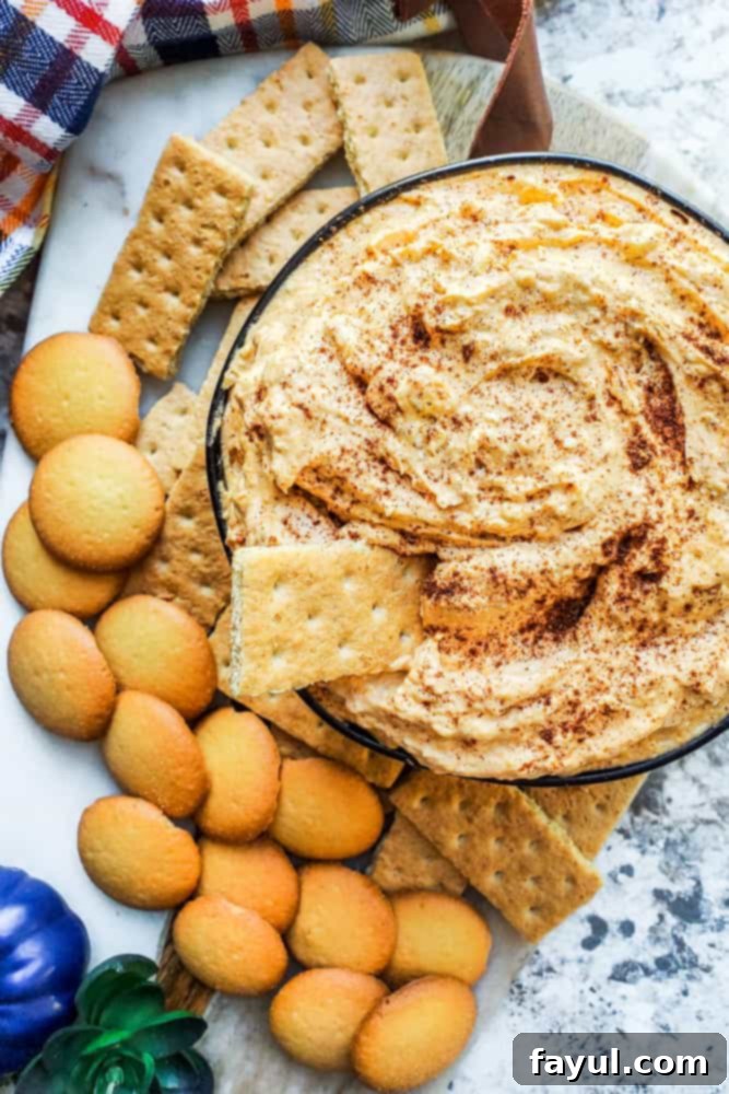 This no bake pumpkin cheesecake dip is ready in only 10 minutes and makes the perfect dessert or appetizer recipe. This delicious dip is filled with cream cheese, cool whip, pumpkin pie spice and brown sugar. Add vanilla wafers, graham crackers or apples to pair with. A perfect easy Fall dessert for Halloween or Thanksgiving. #recipe #appetizer #pumpkin #pumpkinspice #dessert #Fall #nobake #easyrecipe Overhead shot of creamy pumpkin cheesecake dip in a black bowl, garnished with a sprinkle of pumpkin pie spice, on a pristine white counter, surrounded by a variety of dippers like vanilla wafers and graham crackers.