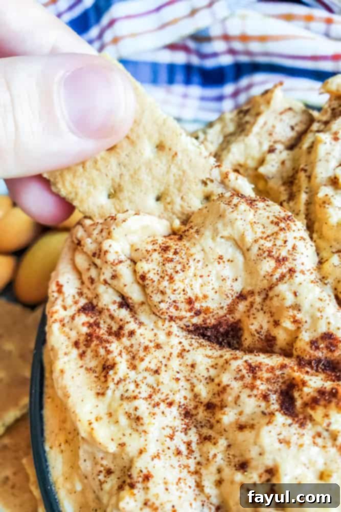A hand reaching into a bowl of pumpkin cheesecake dip with a graham cracker, with a cozy plaid towel subtly in the background, hinting at autumn comfort.