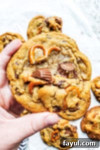 A hand gently holding up a freshly baked peanut butter cup and pretzel cookie, showcasing its rich texture and delicious inclusions.