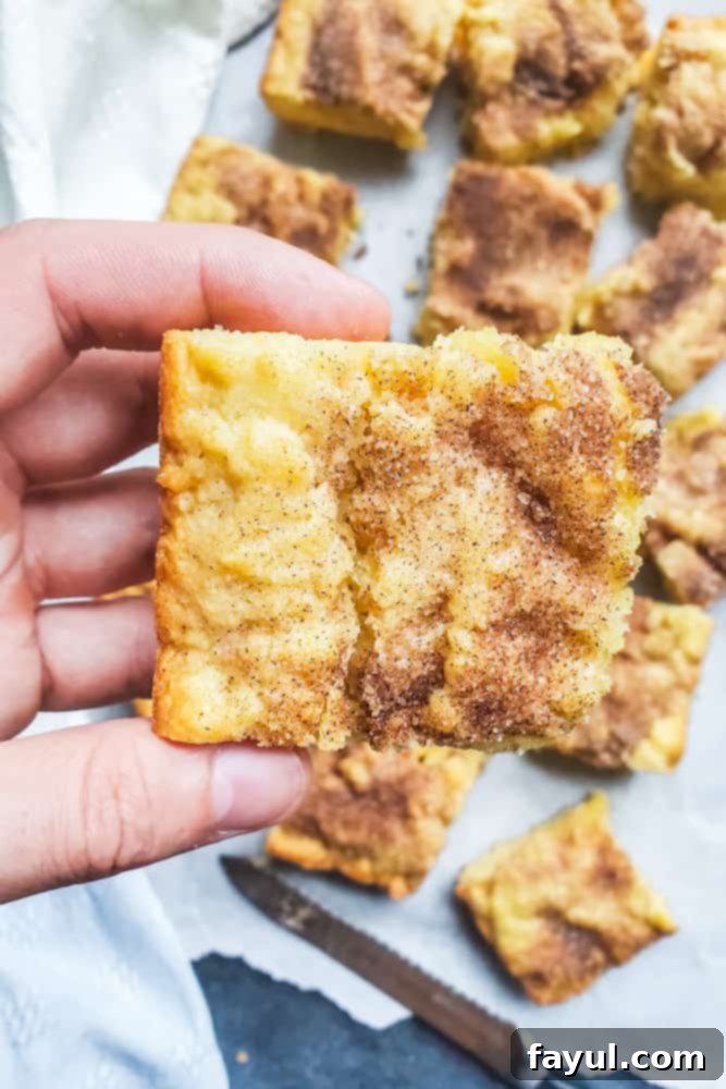 A hand holding a single snickerdoodle blondie, with several other cut blondies arranged on white parchment paper below.